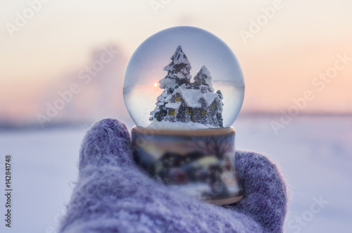 Girl in knitted mittens holding glass ball with firtrees, house and artificial snow with winter landscape at the  background