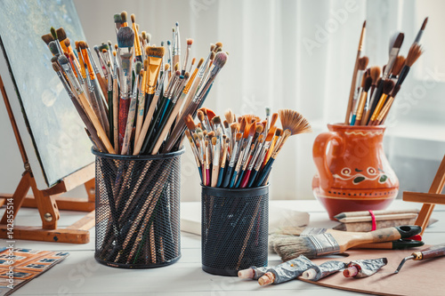 Artistic equipment: easel,  paintbrushes, tubes of paint, palette and paintings on work table in a artist studio. photo