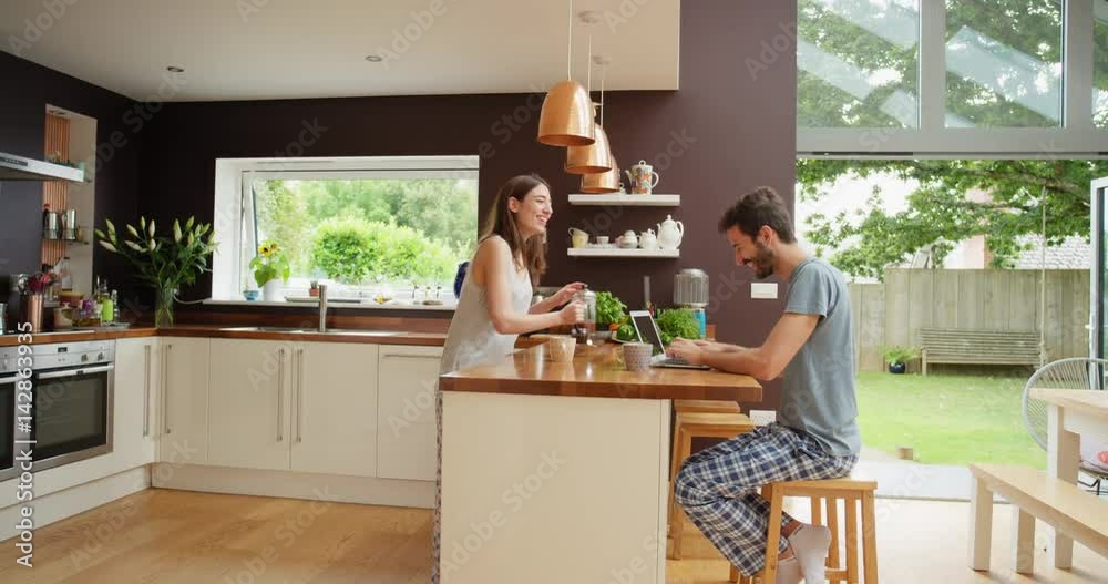 Happy couple at home in kitchen at breakfast using smartphone together browsing online having fun drinking coffee