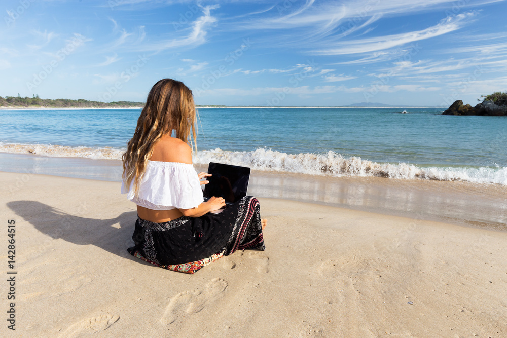 A beautiful young woman sits on the beach beside the sea and crashing waves while working remotely on a laptop.