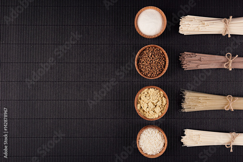 Decorative border of bundles raw noodles with ingredient in wooden bowls on black striped mat background with copy space, top view.