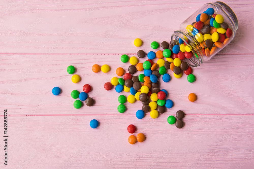 Multicolored candies spilled of a glass jar on a pink wooden background