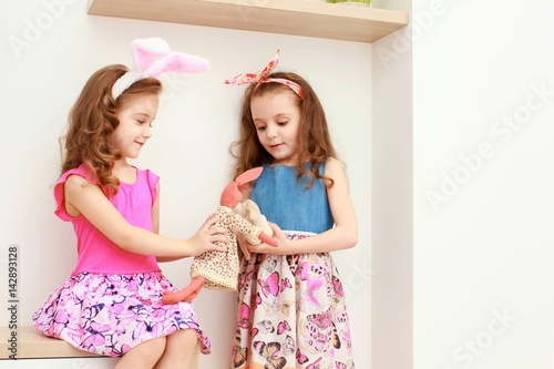Cute little girls with bunny ears and cuddly toy stand near the white wall