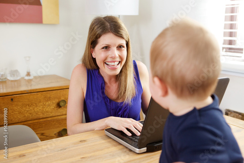 cheerful woman working on laptop and looking at baby son