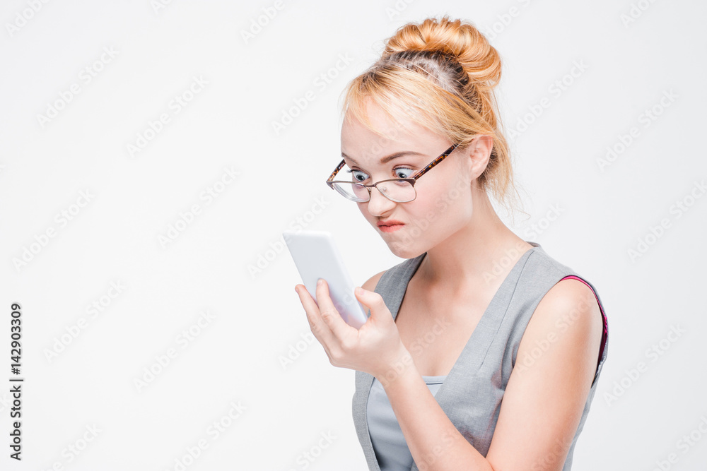 Young woman in glasses with mobile phone looks angry, peeved and irritated. Annoyed and grumpy blonde with smartphone on grey background with copy space. Young woman in glasses with mobile phone looks angry, peeved and irritated. Annoyed and grumpy blonde with smartphone on grey background with copy space.