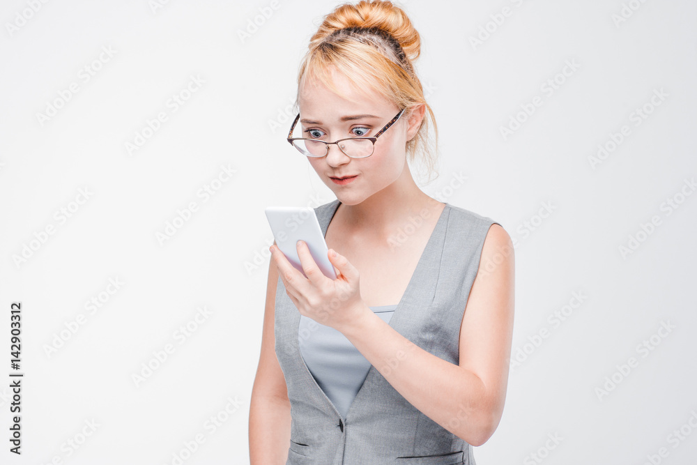 Young woman in glasses with mobile phone looks angry, peeved and irritated. Annoyed and grumpy blonde with smartphone on grey background with copy space. Young woman in glasses with mobile phone looks angry, peeved and irritated. Annoyed and grumpy blonde with smartphone on grey background with copy space.