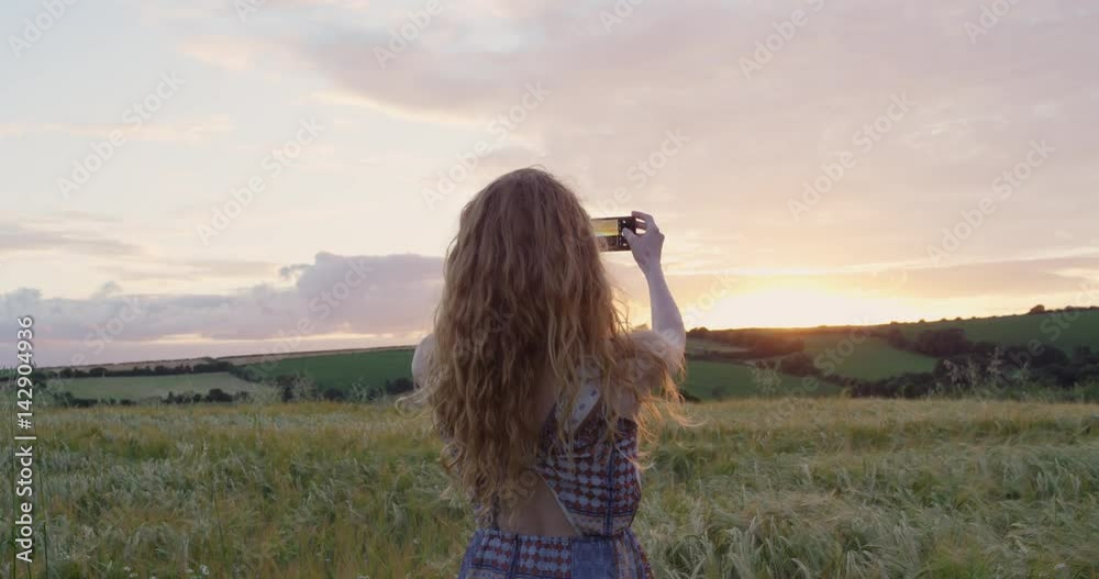 Rear view of attractive red head woman taking photograph wheat field using smartphone capturing epic nature background Cornwall, UK