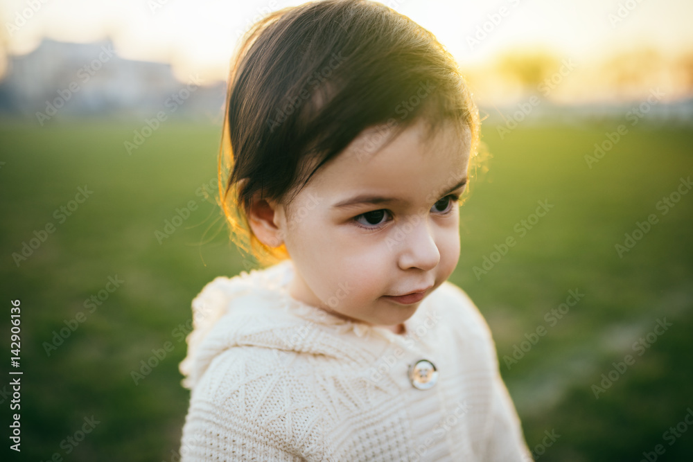 Caucasian girl baby play outdoor. Close up portrait of cute girl toddler looking away on green grass and sunset background. Happy child in outside. Childhood lifestyle. Caucasian girl baby play outdoor. Close up portrait of cute girl toddler looking away on green grass and sunset background. Happy child in outside. Childhood lifestyle.