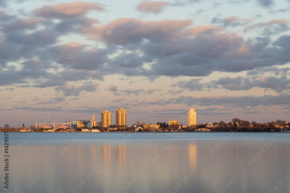 Vibrant cityscape sunset viewed from across a smooth lake reflecting colors, clouds in sky showing motion from wind