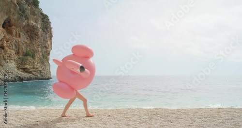 Woman walking on empty beach holding giant inflatable flamingo enjoying summer vacation on tropical beach holiday wearing full one piece swimsuit