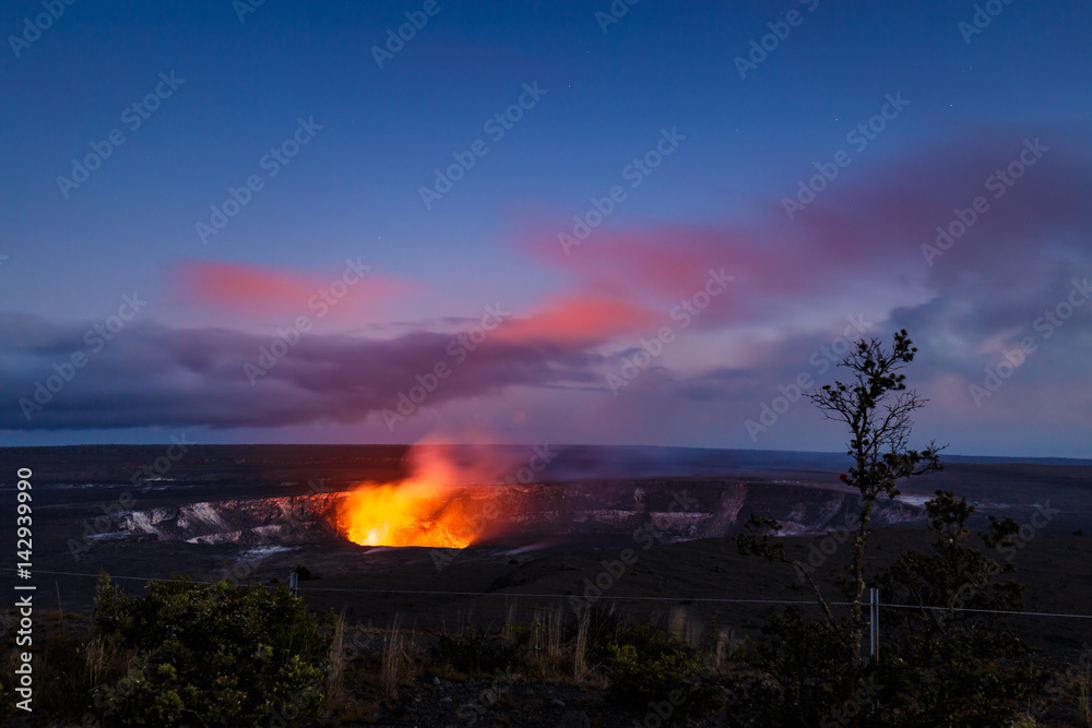 The fiery glow of the lava lake of Kilauea's active crater of Halemaumau illuminates pink clouds at the blue hour. View from Jaggar museum, Volcanoes National Park, Big Island, Hawaii.