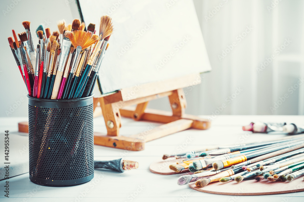 Naklejka premium Artistic equipment: easel, paint brushes, tubes of paint, palette and paintings on work table in a artist studio.