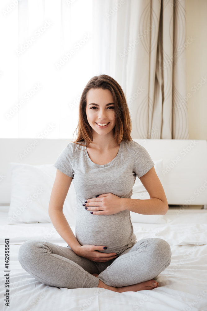 Cheerful pregnant woman sitting on bed with legs crossed Cheerful pregnant woman sitting on bed with legs crossed