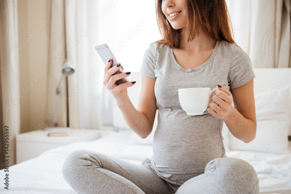 Happy pretty pregnant young woman drinking tea and using cell phone on bed at home Happy pretty pregnant young woman drinking tea and using cell phone on bed at home