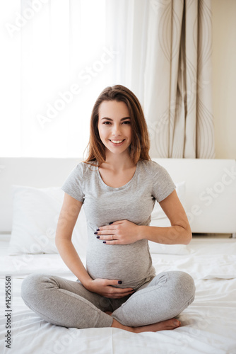 Cheerful pregnant woman sitting on bed with legs crossed