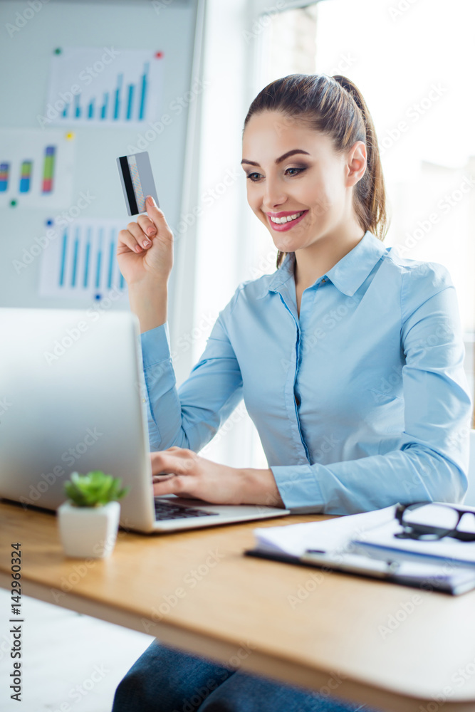 Smiling young charming woman sitting at office holding credit card and doing online shopping Smiling young charming woman sitting at office holding credit card and doing online shopping