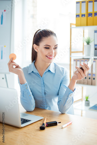 Vertical portrait of charming secretary holding mirror and doing make up at work