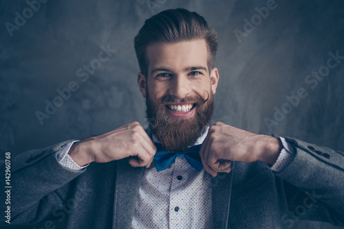 portrait of stylish hipster young man with mustache, beard and beautiful hairstyle make preparation for event party and holding blue bow-tie with toothy smile
