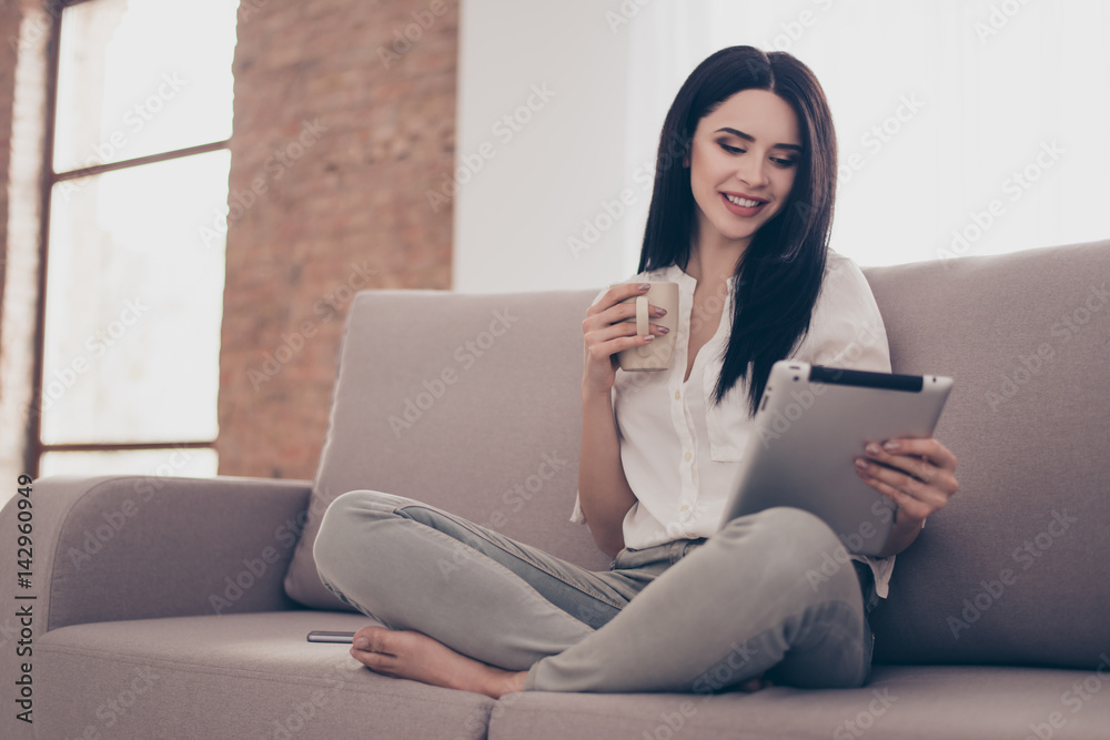 Portrait of smiling charming woman sitting on sofa drinking coffee and reading news from tablet