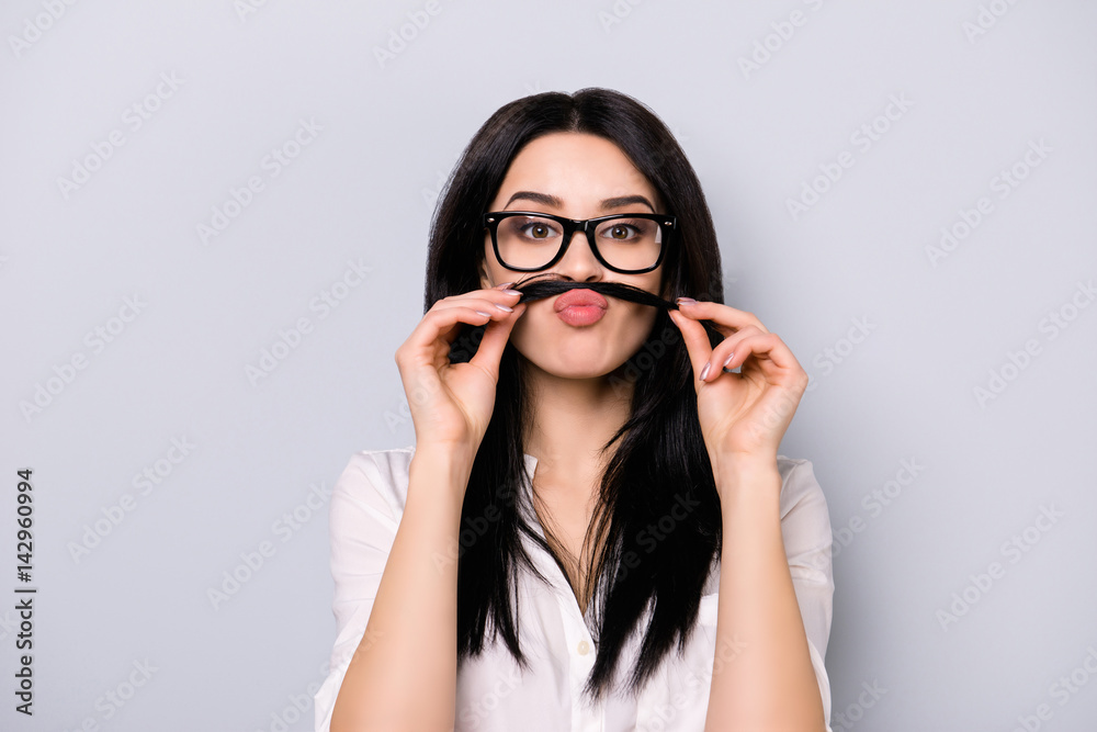 Portrait of funny pretty  young brunette woman with eyeglasses holding hair above lips like have mustaches while standing on gray background