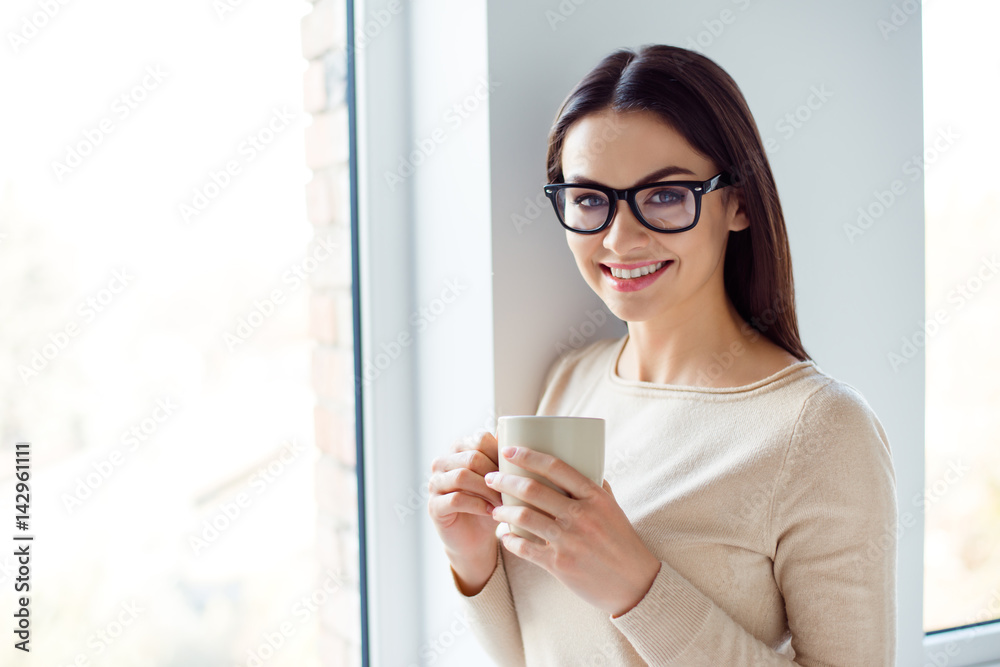 Young smart successful smiling woman in glasses having break, holding cup and drink coffe while standing near window. Young smart successful smiling woman in glasses having break, holding cup and drink coffe while standing near window.