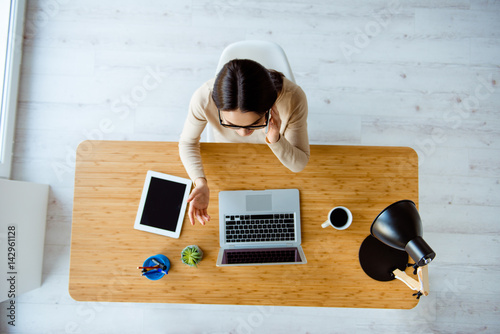 Top-view of young successful woman in glasses sitting at table with laptop, cup of coffee and tablet in office while talk on the phone.