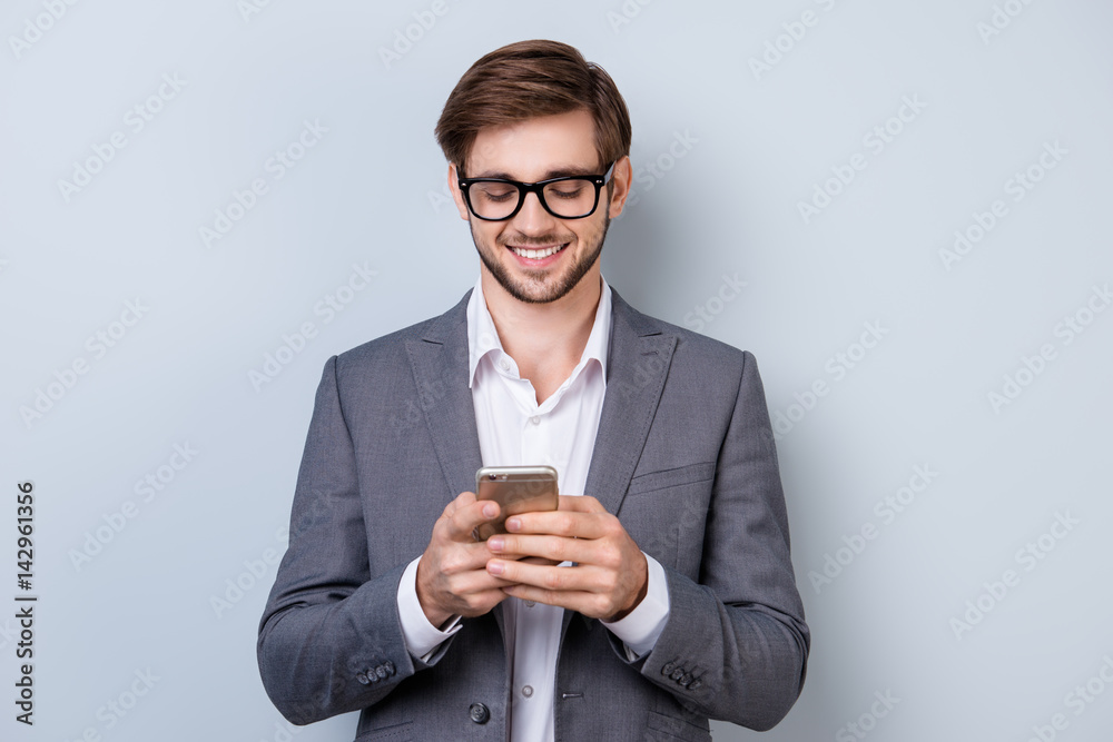 Portrait of successful handsome young man in formal wear with beaming smile, bristle and glasses holding smartphone and read sms while standing on gray background Portrait of successful handsome young man in formal wear with beaming smile, bristle and glasses holding smartphone and read sms while standing on gray background