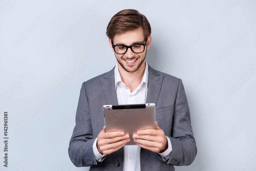Happy young man in formal wear with smile holds digital tablet and works with it while standing on gray background Happy young man in formal wear with smile holds digital tablet and works with it while standing on gray background