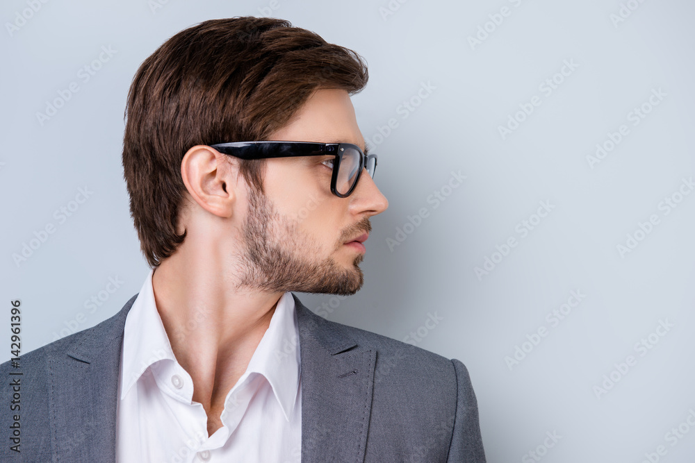 Side-view portrait of young successful serious man with bristle in glasses and wearing suit standing on gray background Side-view portrait of young successful serious man with bristle in glasses and wearing suit standing on gray background