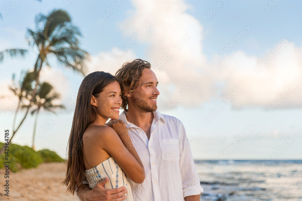 Obraz premium Relaxing casual couple watching sunset together on Waiohai beach, Poipu in Kauai, Hawaii. Beautiful multiracial couple relaxing on summer holidays.