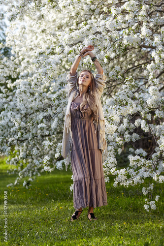 Beautiful young woman in long dress boho style on green grass under apple tree in blossom in garden