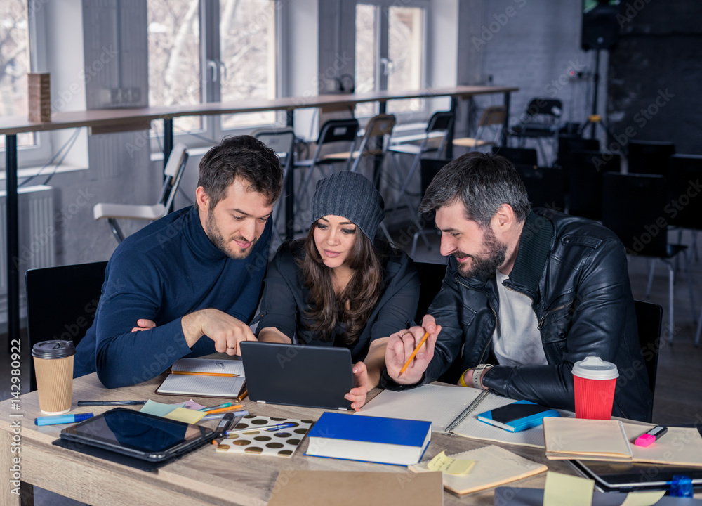 Group of modern people watching project on tablet in the office. Working environment with laptop, coffee, notepads and stationery. They are looking at the screen. Business team concept. Group of modern people watching project on tablet in the office. Working environment with laptop, coffee, notepads and stationery. They are looking at the screen. Business team concept.