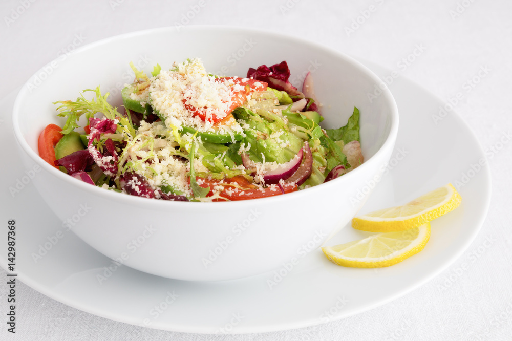 a bowl of green leafs, tomato, avocado, and red onion salad with oil and vinegar dressing and grated cheese decorated with lemon slices