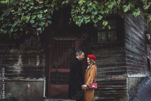 European couple at street, autumn look, casual style. Red beret and coat. Wooden facade of old building photo