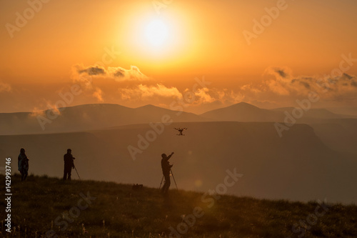 photographers at sunrise high in the mountains launching quadcopter, soft warm background