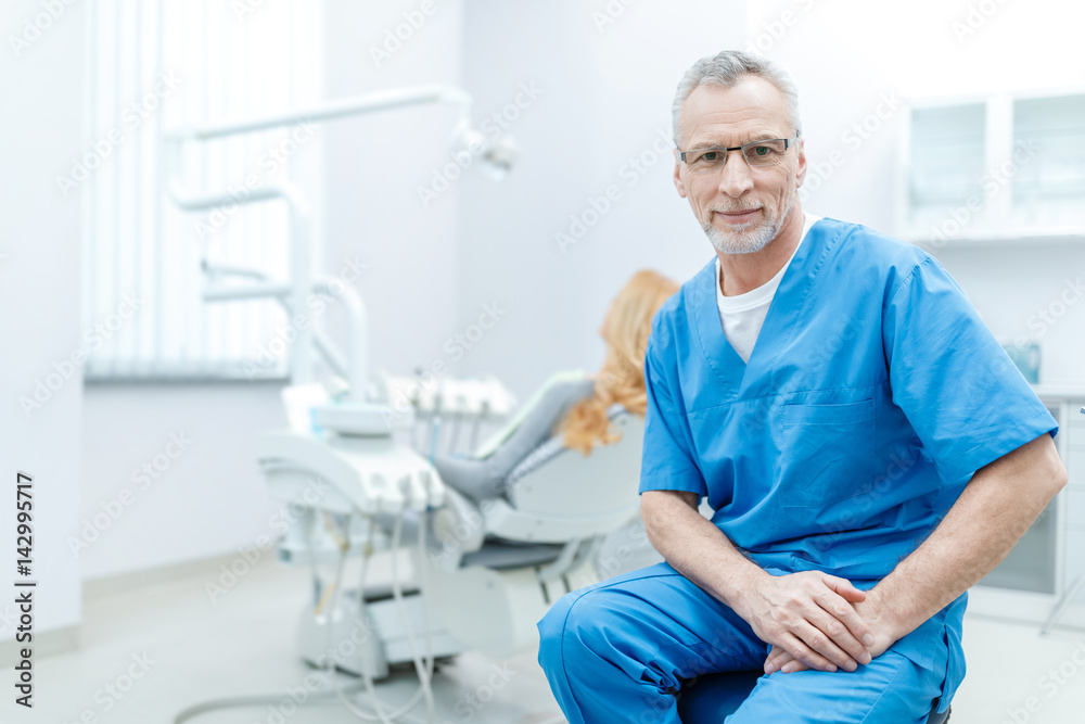 senior dentist in uniform sitting in dental clinic with patient behind senior dentist in uniform sitting in dental clinic with patient behind