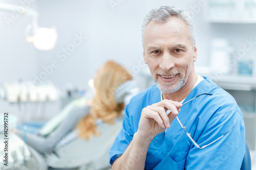 senior smiling dentist in uniform in dental clinic with patient behind