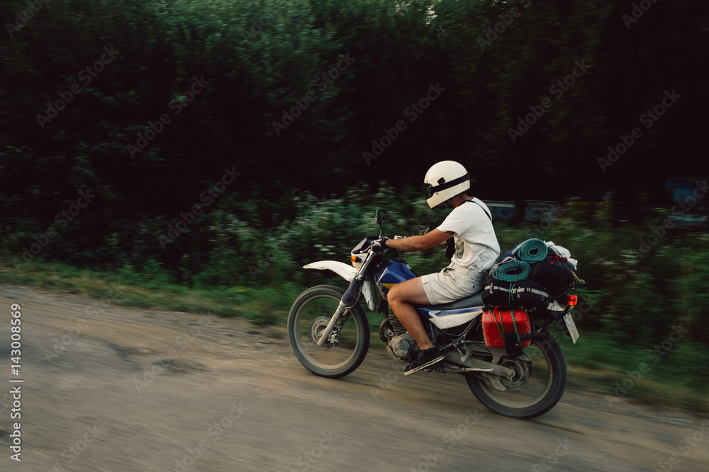 Young man riding motocycle scrambler on pothole filled road kicking up dust loaded with camping gear in the green wilderness off the grid.