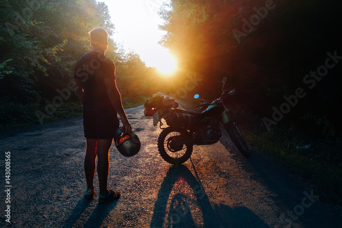 Young man holding helmet next to motorcycle scrambler loaded with camping gear for a moto tour staring into the sunset. © onewithahalf