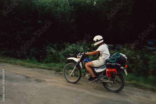 Young man riding motocycle scrambler on pothole filled road kicking up dust loaded with camping gear in the green wilderness off the grid.
