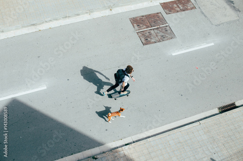 Young hipster with curly hair longboarding commuting with dog running behind on the concrete urban street white light tattoos shadow shot from above.