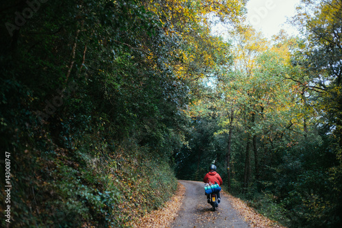 Selective focus shot of the motorcyclist biker riding a narrow rural road in the autumn forest surrounded by beautiful scenery near the camping ground