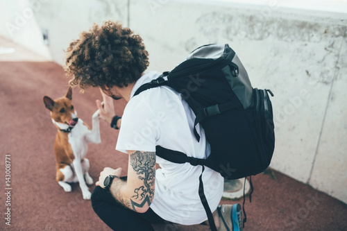 Young trendy hipster with tattoos crazy curly hair with his best friend basenji dog puppy give each other high five while the owner wears a backpack