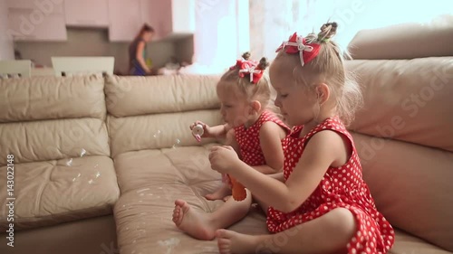 Two little twin sisters in charming red dresses are blowing soap bubbles cheerfully while sitting in the room barefoot. photo