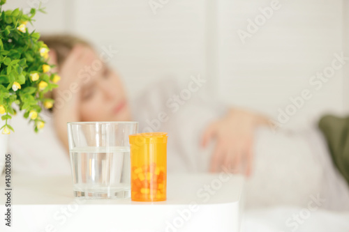 Glass of water, box with pills on table and blurred woman suffering from headache on background