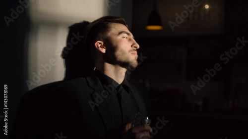 The serious man with black hair and suit is drinking light beer and standing near the wall of the dack pub. The close-up side portrait. photo