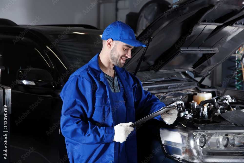 Mechanic standing in front of open car hood with clipboard Mechanic standing in front of open car hood with clipboard