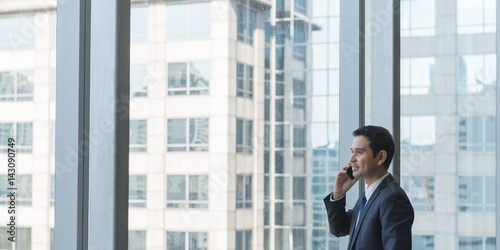 Mature and confident business executive looking looking out of large windows at a view of the city below, from the top floor of an office building, while talking on his mobile phone - panoramic banner