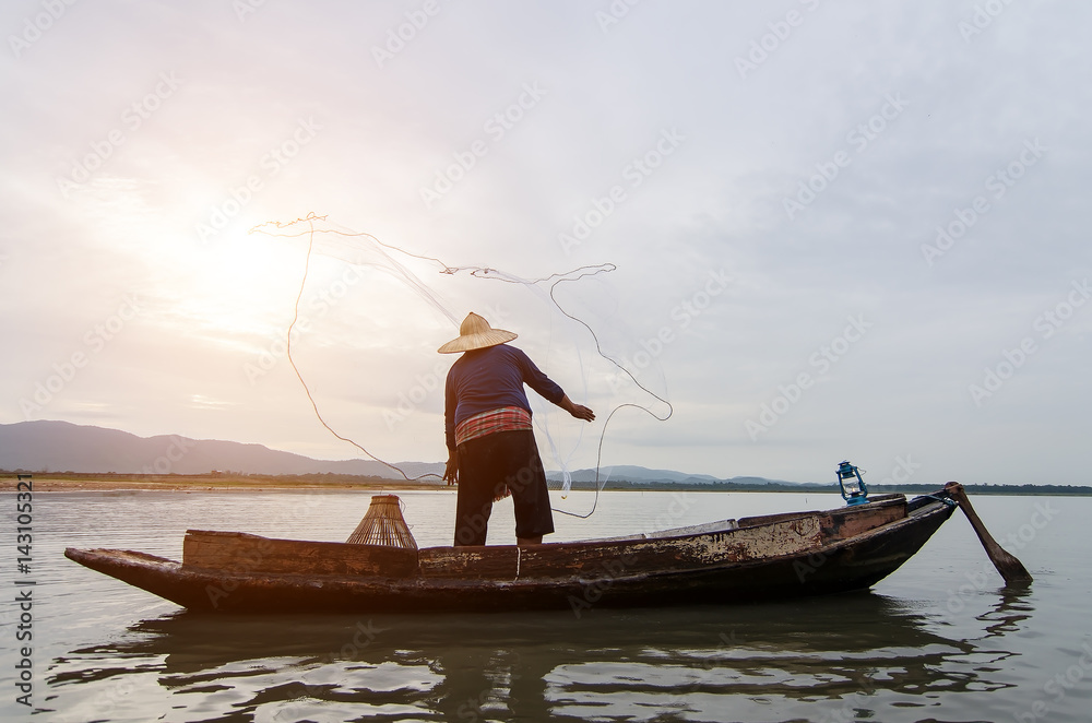 Fototapeta Asian fisherman on wooden boat casting a net for catching freshwater fish in nature river in the early morning before sunrise