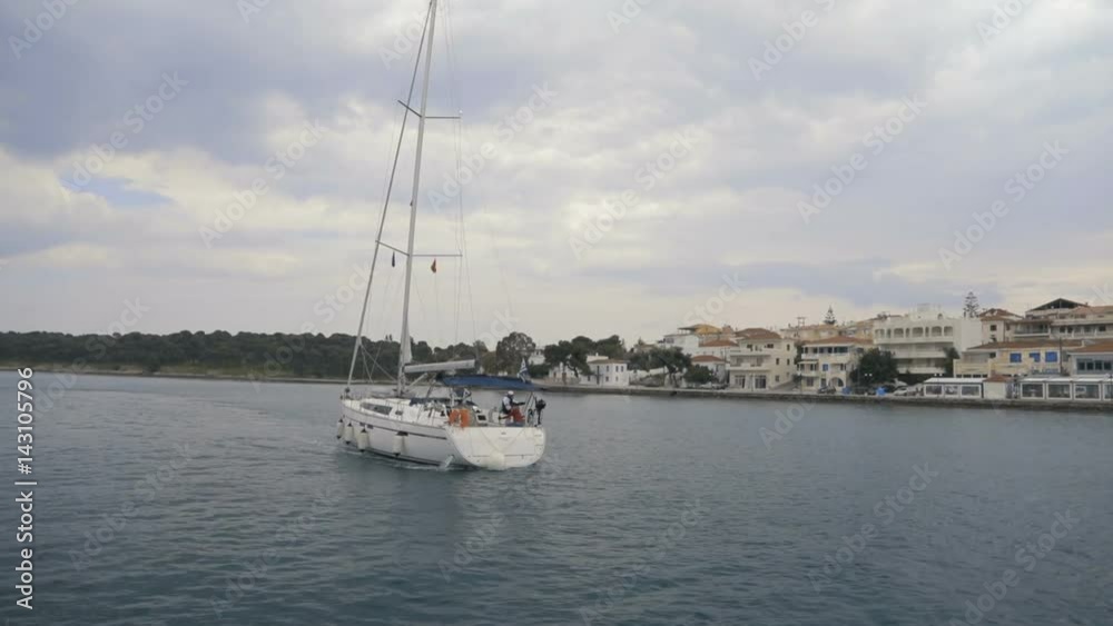 White sail boat with mast heading to the open sea. Ship passing island with small town with villas at the seafront. White sail boat with mast heading to the open sea. Ship passing island with small town with villas at the seafront.